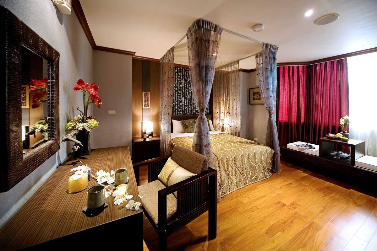 A bedroom with a four-poster bed, a wooden desk with a chair, a bedside table, and a large mirror.