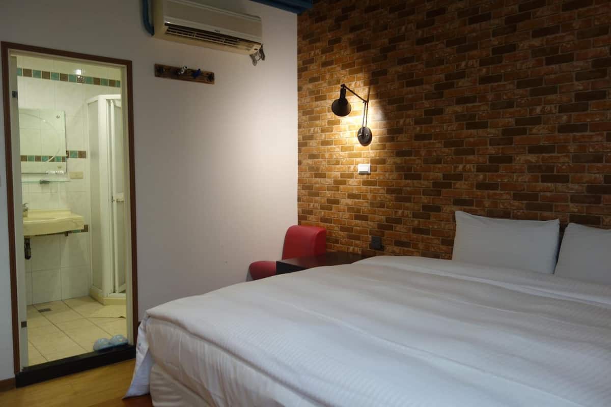 A bedroom with a large bed, a red chair, a brick wall, and a bathroom visible through a glass door.