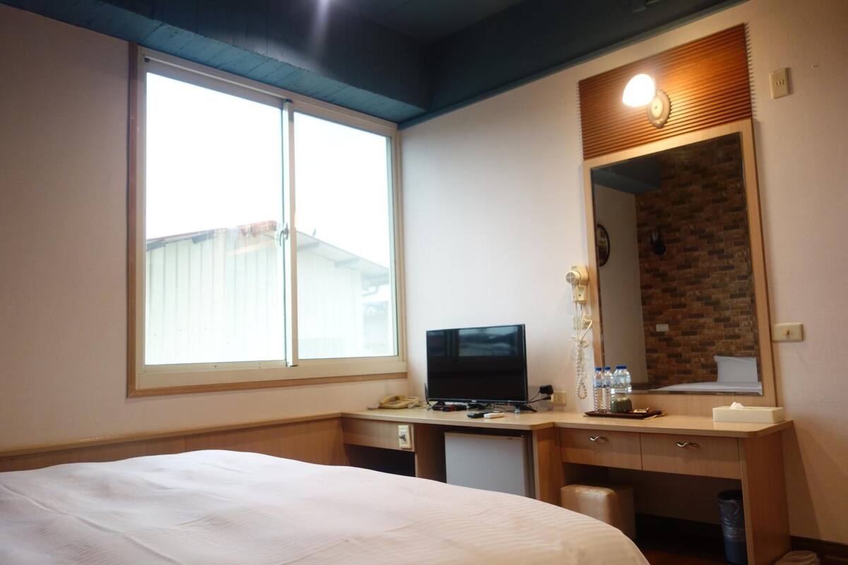 A hotel room with a bed, a desk with a computer, a mirror, and a window.