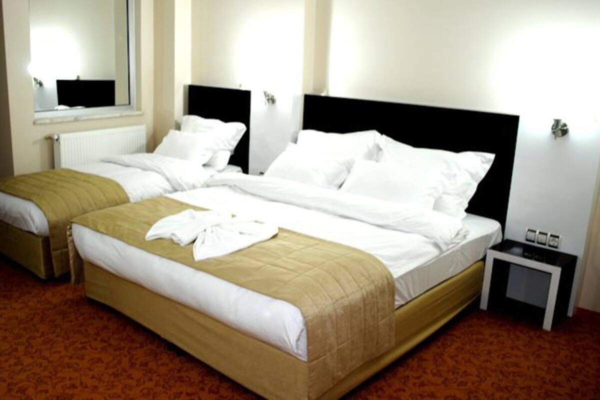 A double bed with white linens, a bedside table with a lamp, and a window with blinds.
