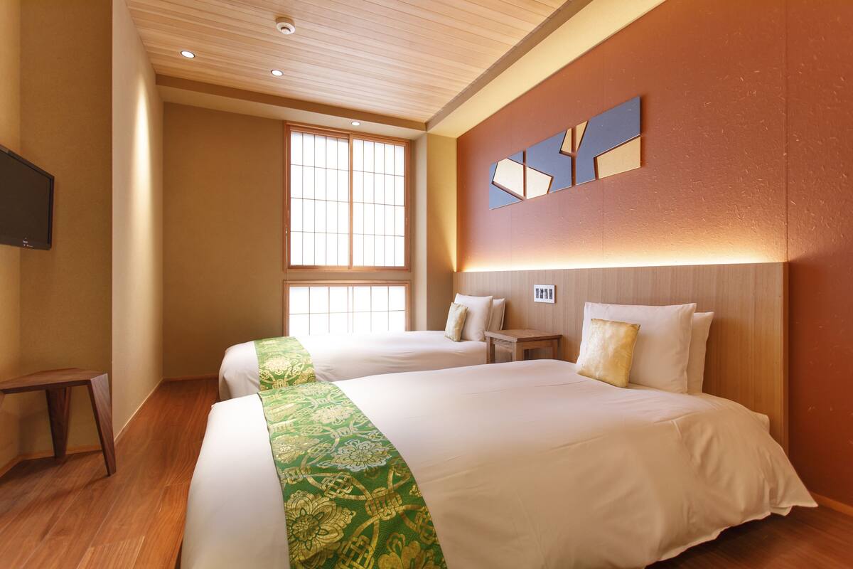Habitación de hotel con dos camas, suelo de madera, un ventanal grande y un televisor de pantalla plana montado en la pared.
