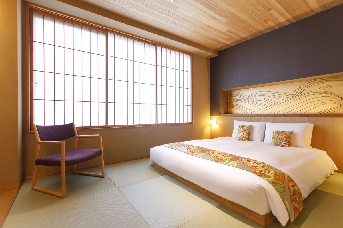 Habitación de hotel con suelo de tatami, cama con cabecera decorativa, una silla y un ventanal grande con marcos de madera.