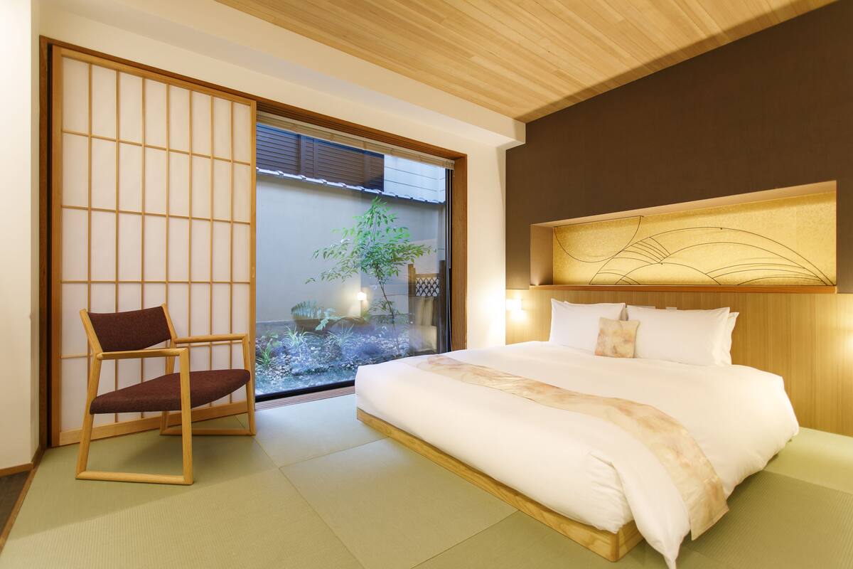 Habitación de hotel con una cama grande, una silla de madera, un panel deslizante shoji y vistas de un jardín a través de un vidrio.