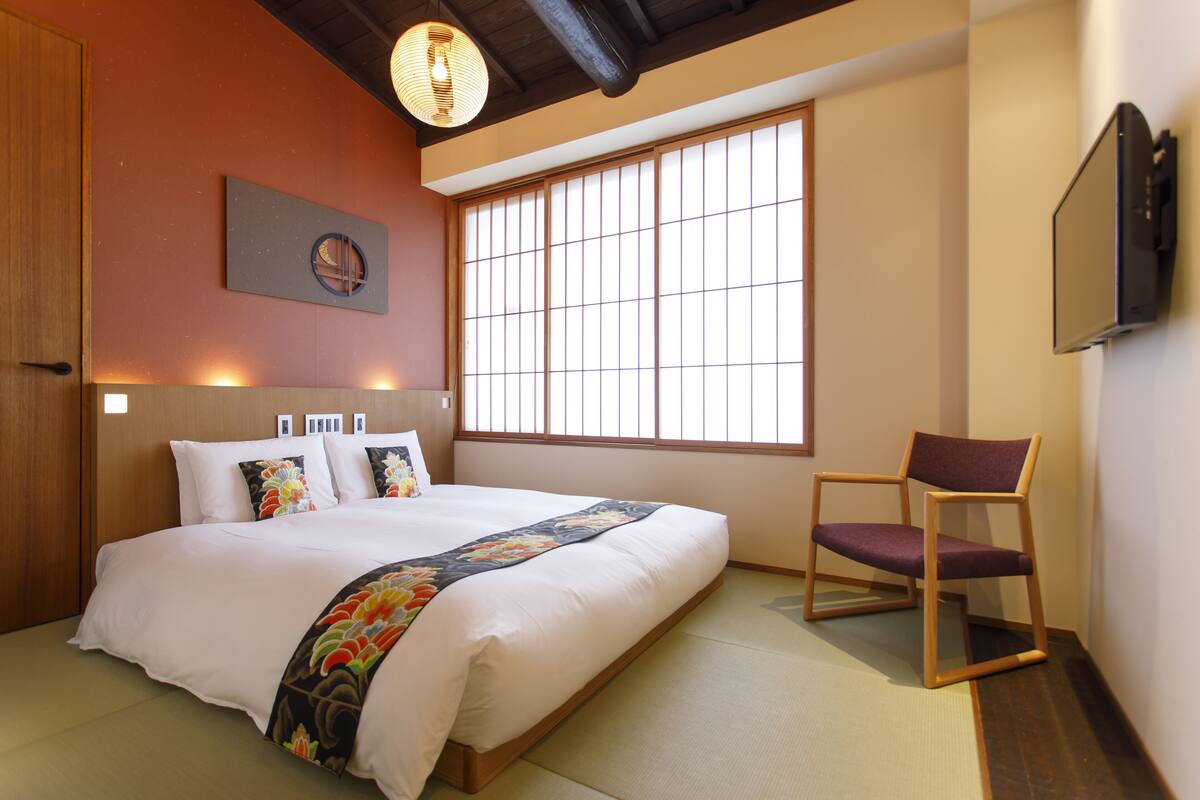 Un dormitorio japonés tradicional con suelo de tatami, un ventanal con puertas correderas shoji, una cama con ropa de cama blanca, una silla de madera y un televisor montado.