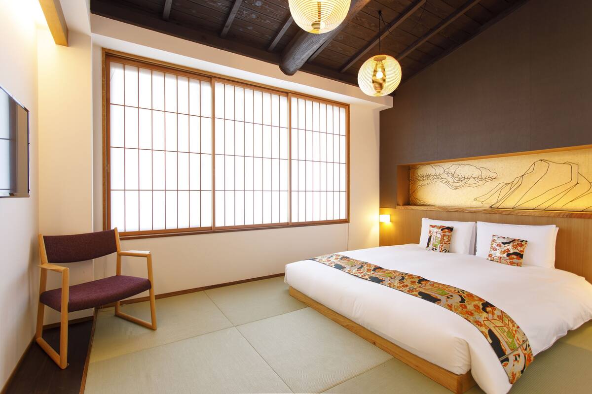 Un dormitorio de estilo japonés tradicional con suelo de tatami, una cama de madera baja, una ventana con celosías shoji y un colgante de luz.