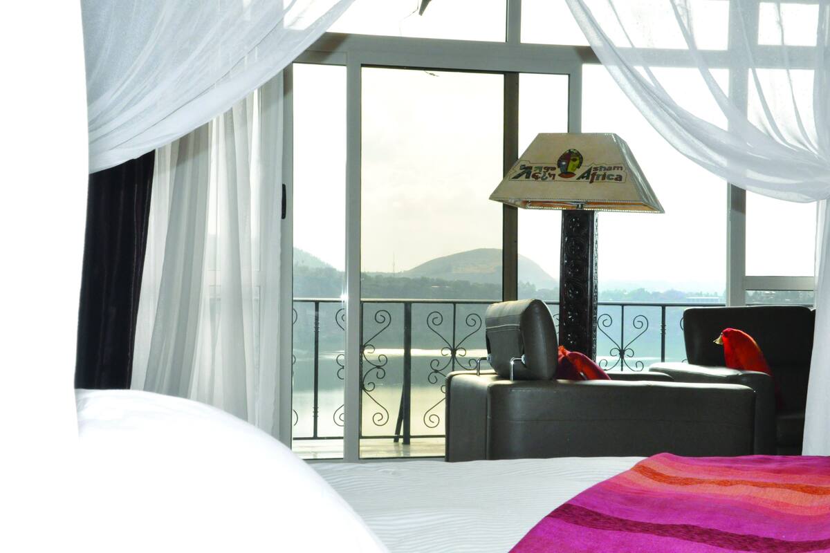 A hotel room with a bed, a chair, a table, and a view of a body of water and mountains.
