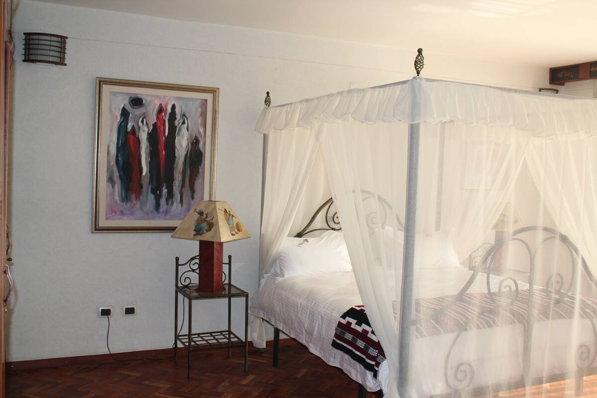A bedroom with a four-poster bed, a bedside table with a lamp, and a painting on the wall.