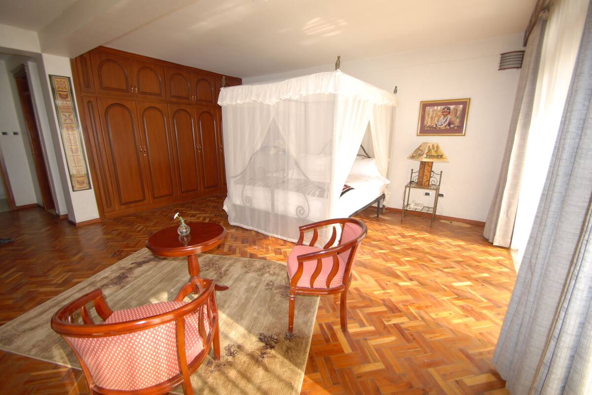 A room with a wooden floor, a bed with a canopy, a chair, a small table, and a lamp.