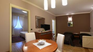 A hotel room with a wooden dining table, white chairs, a sofa, a TV, and a mirror.