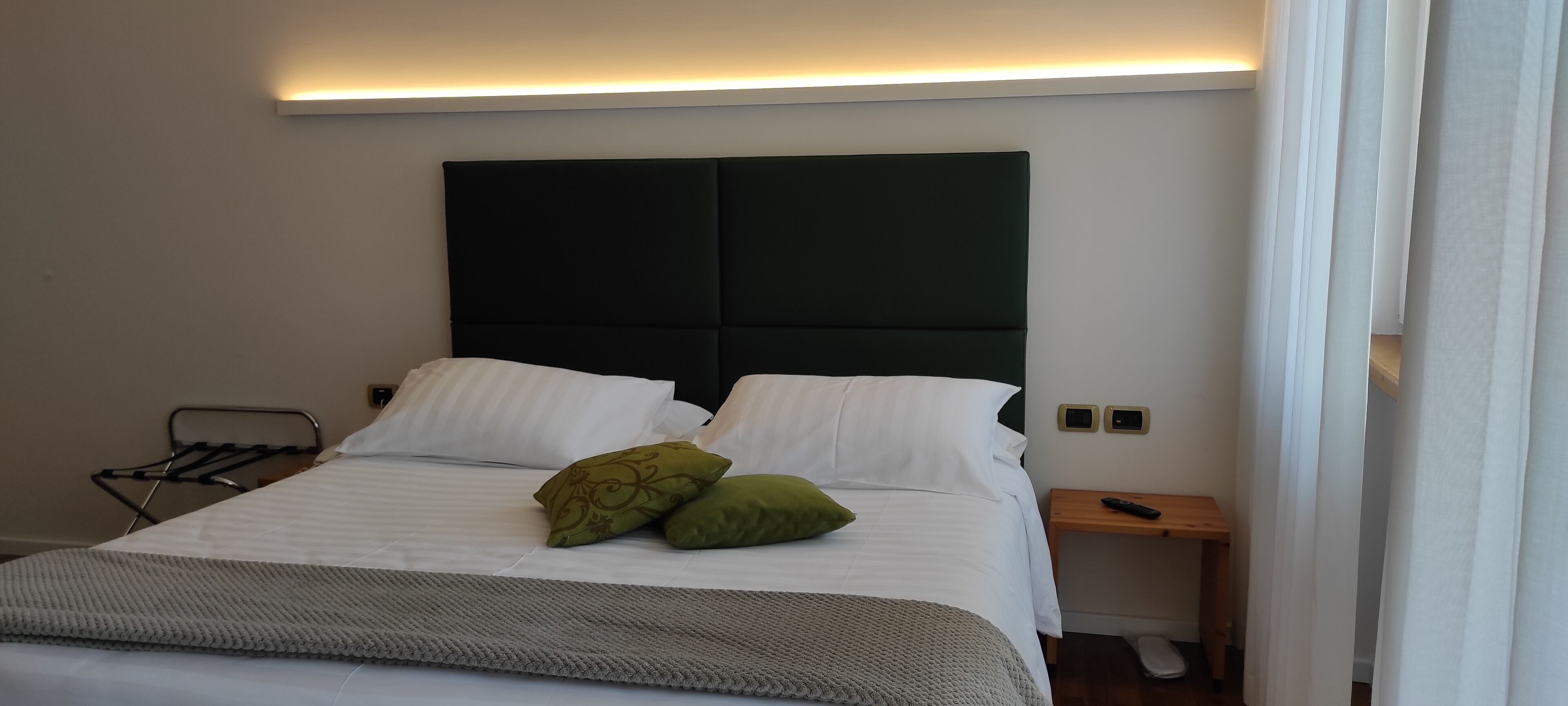 Una cama bien hecha con sábanas blancas y dos almohadas verdes, una mesita de noche con un control remoto y una lámpara de pared.