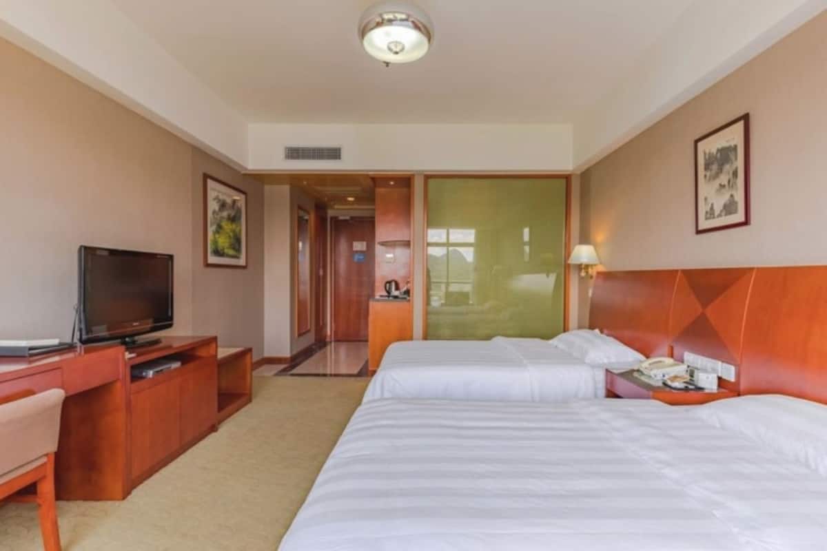 Habitación de hotel con dos camas, televisión, escritorio y baño visible a través de una puerta de cristal.