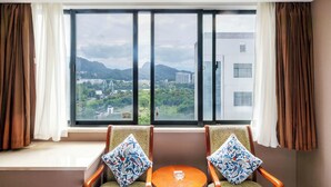 A hotel room with a large window, two armchairs, a small round table, and a view of a cityscape and mountains. - Huangshan Tiandu International Hotel (Huangshan)