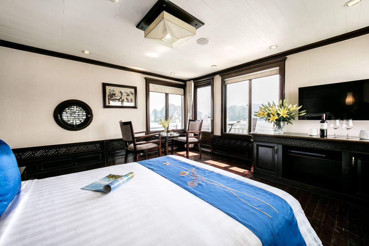 A hotel room with a large bed, a dining table with chairs, a TV, and a view of the water.