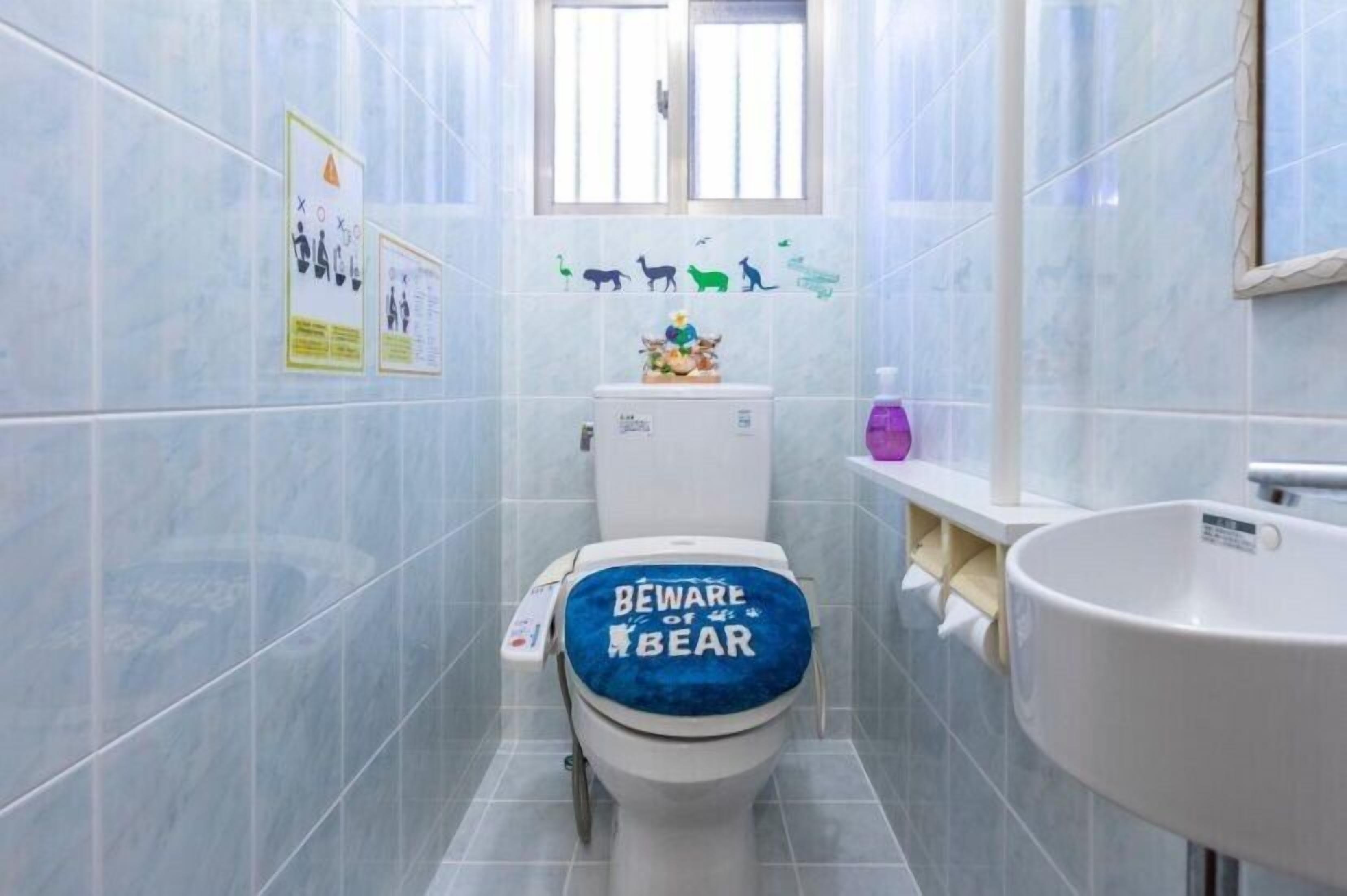 A bathroom with a toilet covered by a blue seat, a sink, and a window above the toilet.