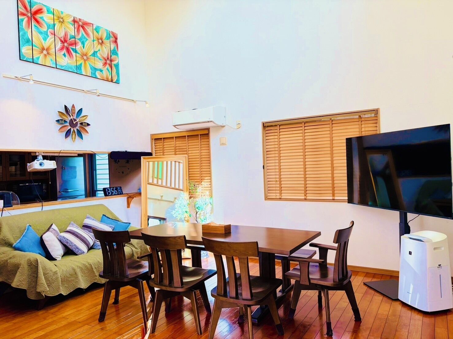 A cozy living room with a wooden floor, a green sofa, a dining table with chairs, a large TV, and a colorful abstract painting on the wall.