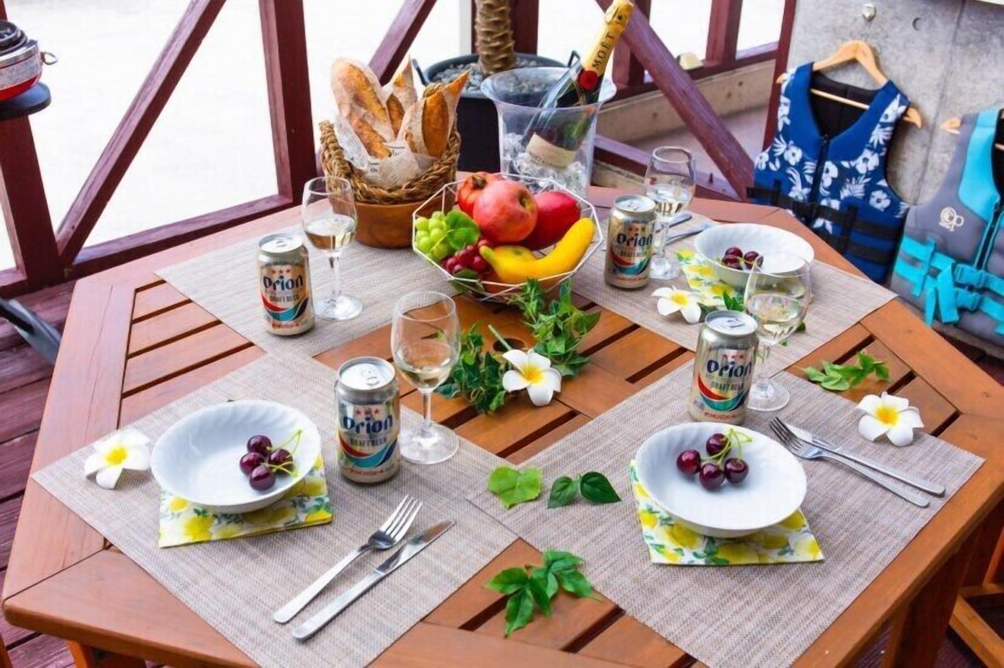 Een gedekte tafel met drankjes, fruit en bloemen, met een champagnefles en een broodmand in de achtergrond.