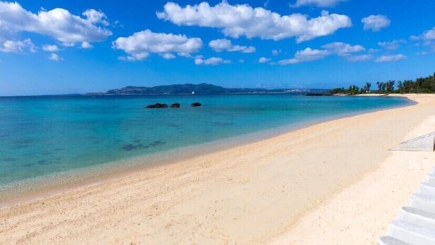 Una playa con agua turquesa cristalina, una orilla arenosa y una cadena montañosa distante bajo un cielo azul con nubes dispersas.
