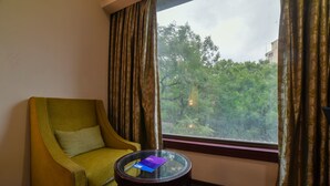 A room with a green armchair, a glass table with a book, and a window with curtains. - Hotel Mint Oodles (New Delhi)