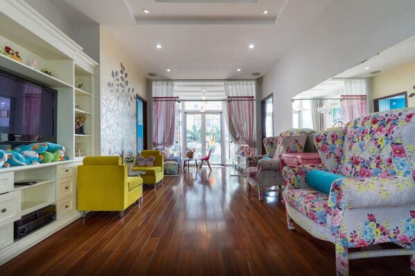 Un salón amplio con suelo de madera, un sillón amarillo, un sofá con estampado floral y un televisor sobre un mueble blanco.