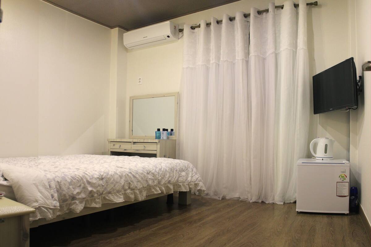 A hotel room with a bed, a small bedside table, a mirror, a TV mounted on the wall, a white radiator, and a white curtain.