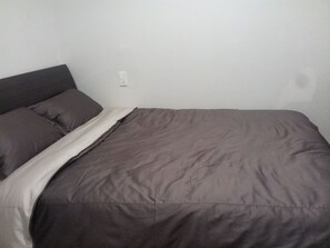 A bed with a dark brown mattress and pillows, a white blanket, and a plain white wall with a light switch.