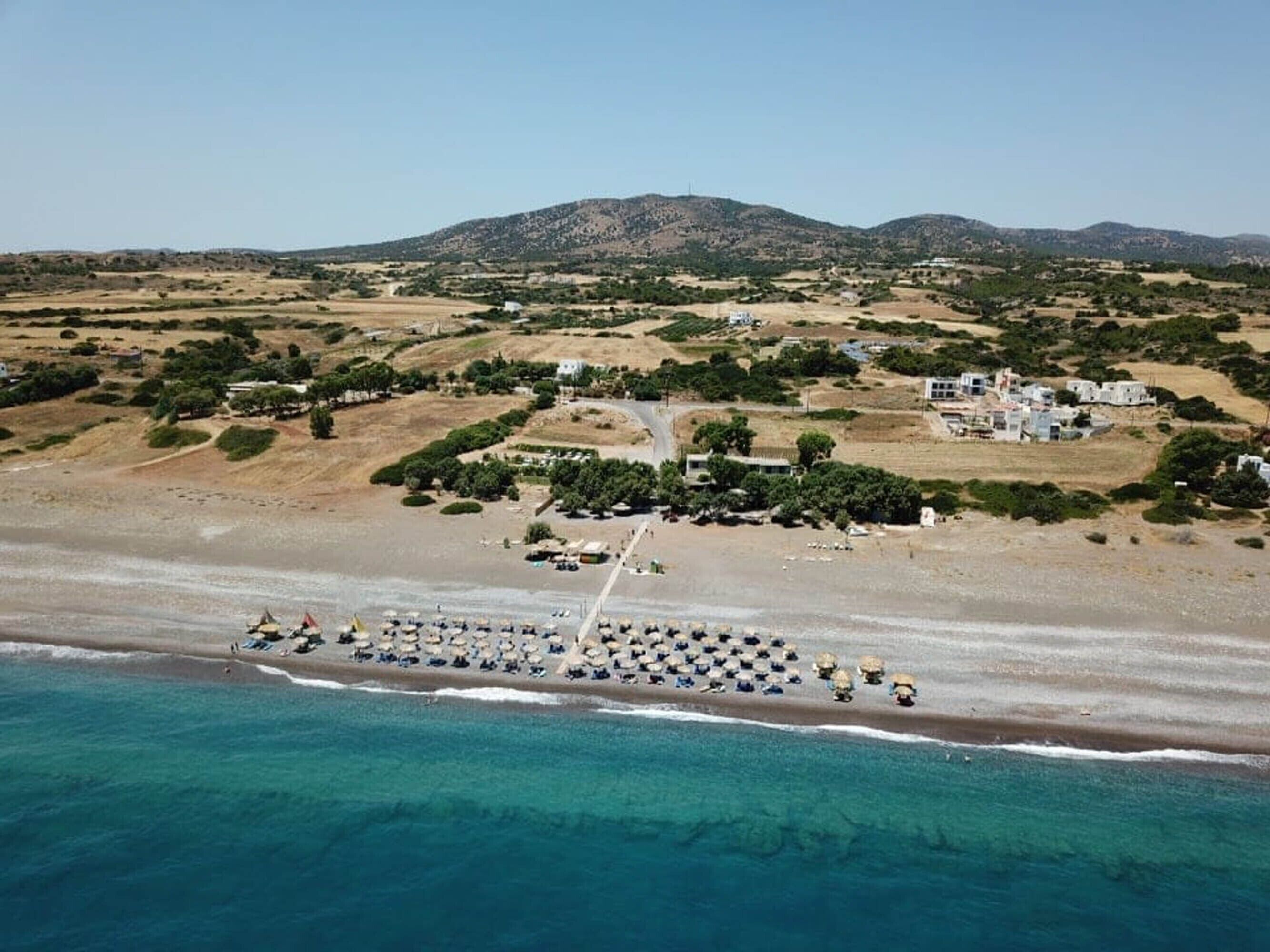 An aerial view of a coastal area with a beach, a row of beach umbrellas, and a cluster of buildings in the background.
