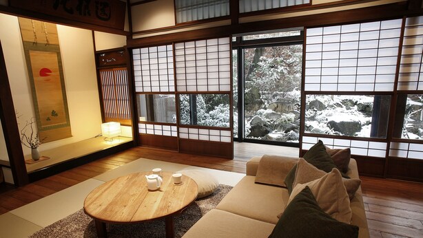 Una habitación tradicional japonesa con suelo de tatami, una mesa de madera baja con tazas, un sofá con cojines y grandes shoji con vistas a un jardín nevado.