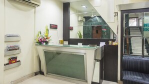 Un área de recepción con un mostrador de vidrio, unidades de aire acondicionado, un reloj digital y una escalera que conduce a un nivel superior.