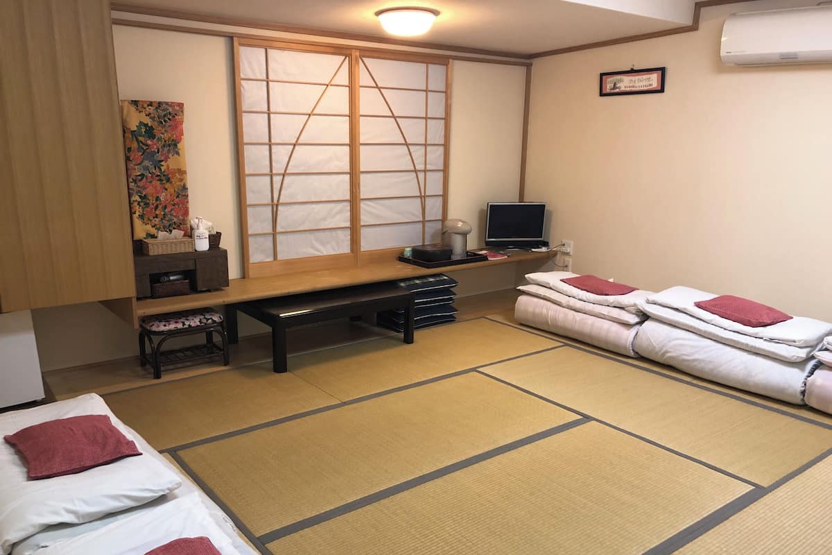 Ein Zimmer mit Tatami-Boden, einem Futon, einem Schreibtisch mit Computer, einem Fernseher und einer traditionellen japanischen Schiebetür.