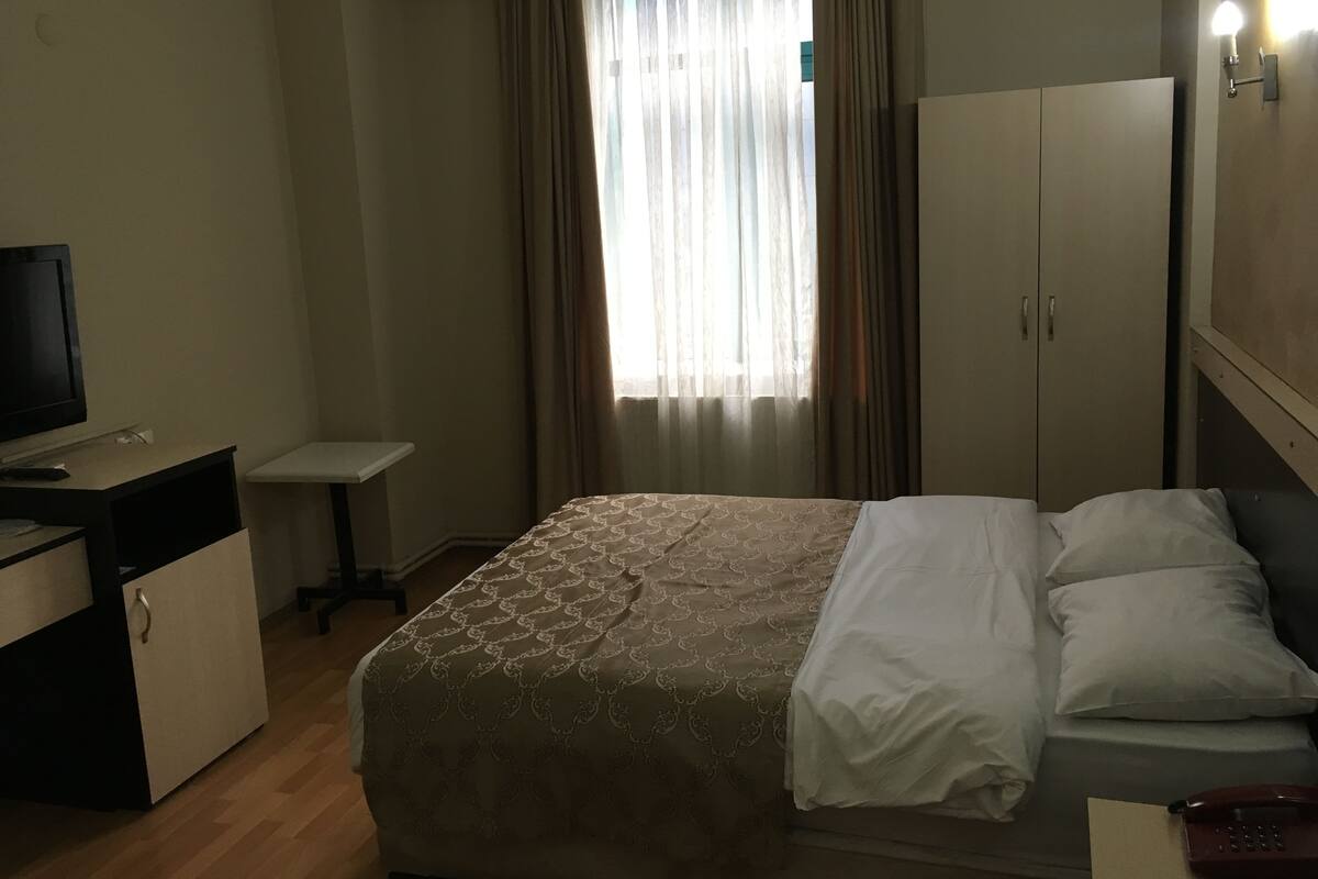 A hotel room with a bed, a desk with a television, a wardrobe, and a window with curtains.