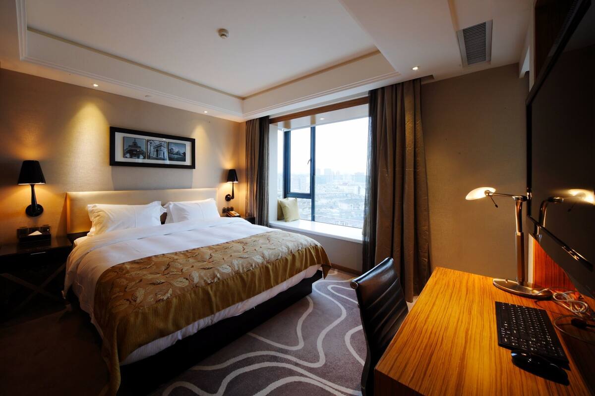 Ein Hotelzimmer mit einem großen Bett, einem Schreibtisch mit Computer und einem Fenster mit Blick auf die Stadt.
