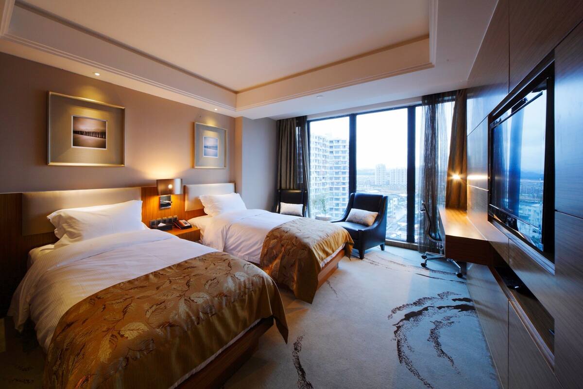 Ein Hotelzimmer mit zwei Betten, einem Schreibtisch, einem Stuhl und großen Fenstern mit Blick auf die Stadt.