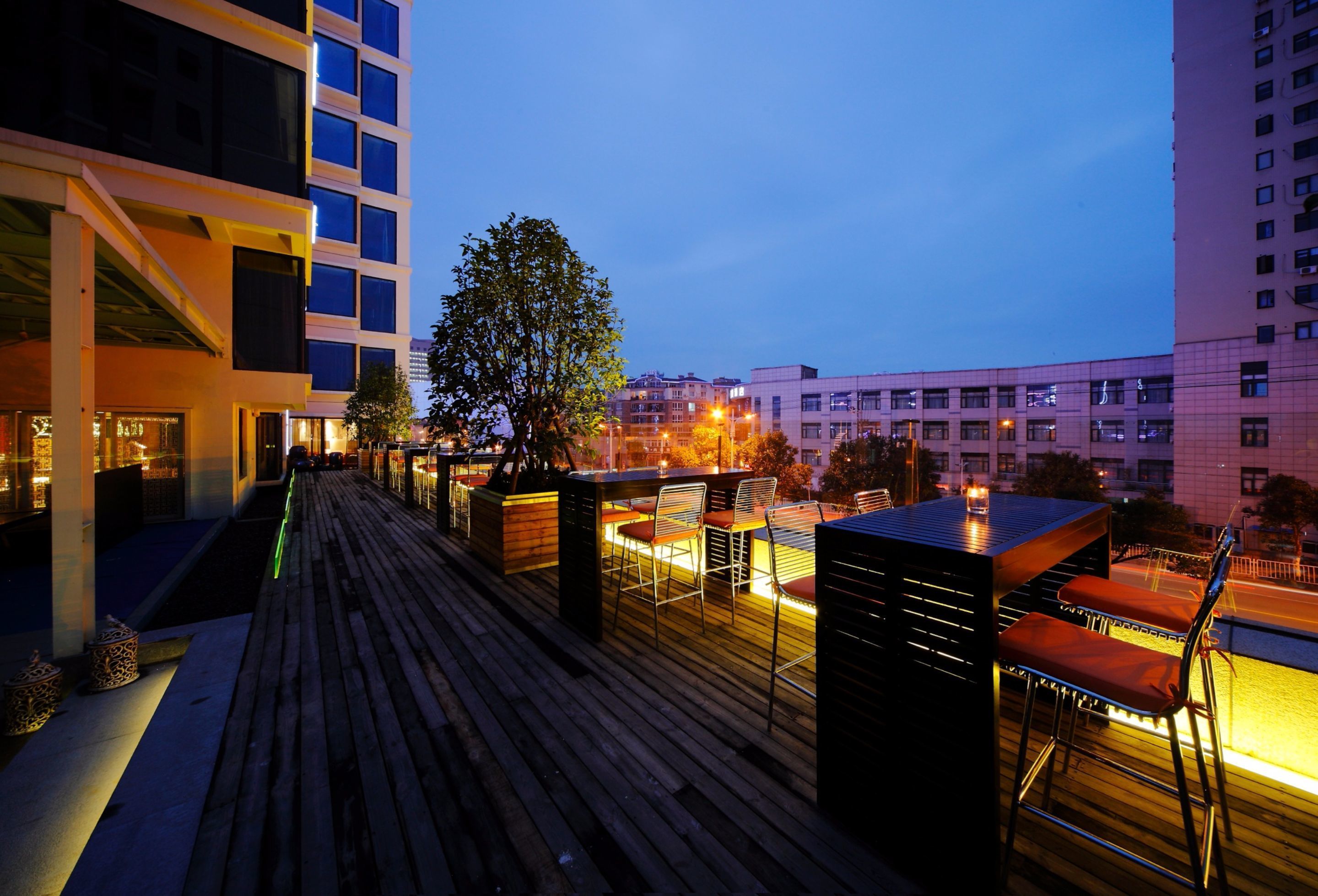 Un bar situé sur un toit, avec une terrasse en bois, des sièges en plein air et une vue sur la ville au crépuscule.