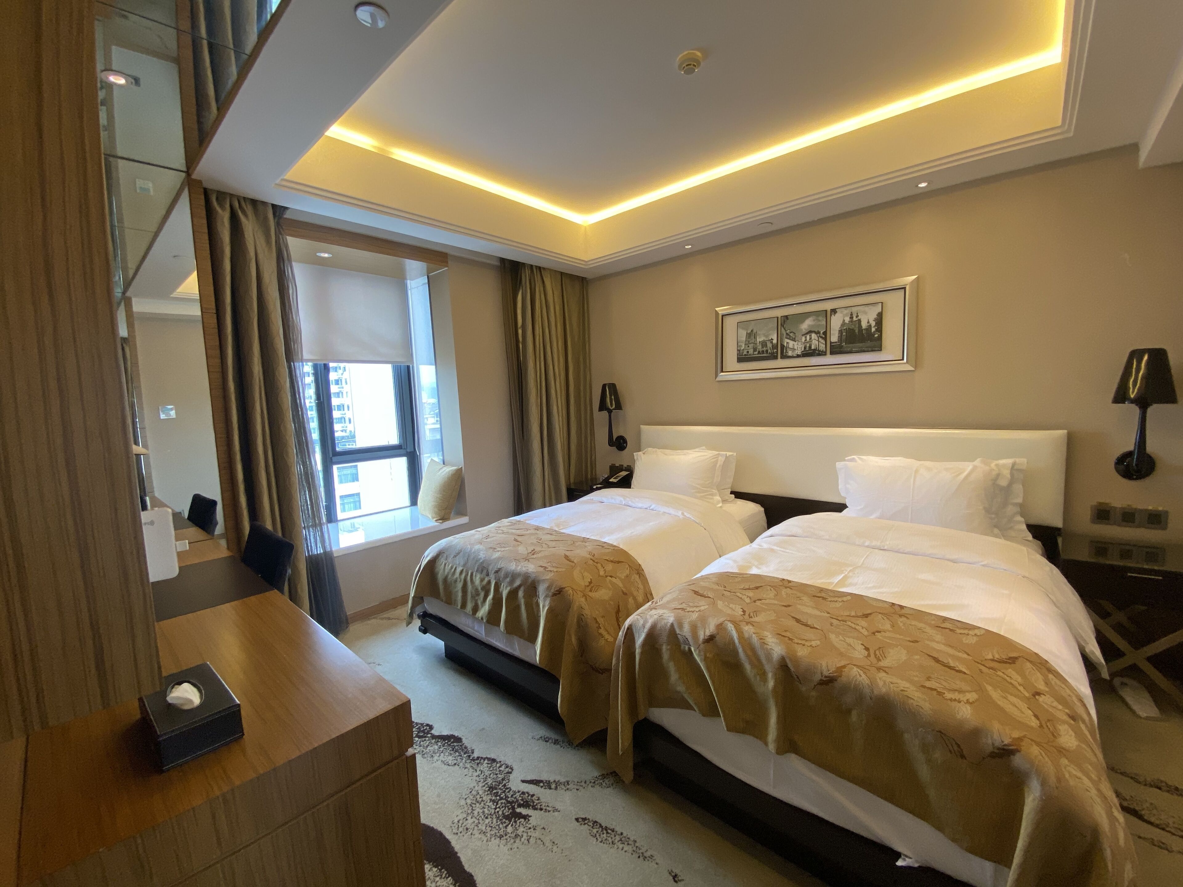 Une chambre d’hôtel avec deux lits, une table de chevet, un miroir, une fenêtre avec des rideaux, un tableau accroché au mur et un luminaire au plafond.