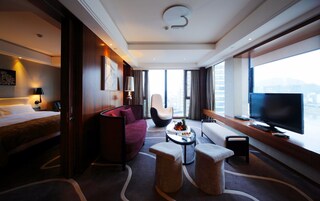 Una habitación de hotel moderna con una cama, un sillón rojo, una mesita de centro redonda con fruta, un televisor y vistas a la ciudad.