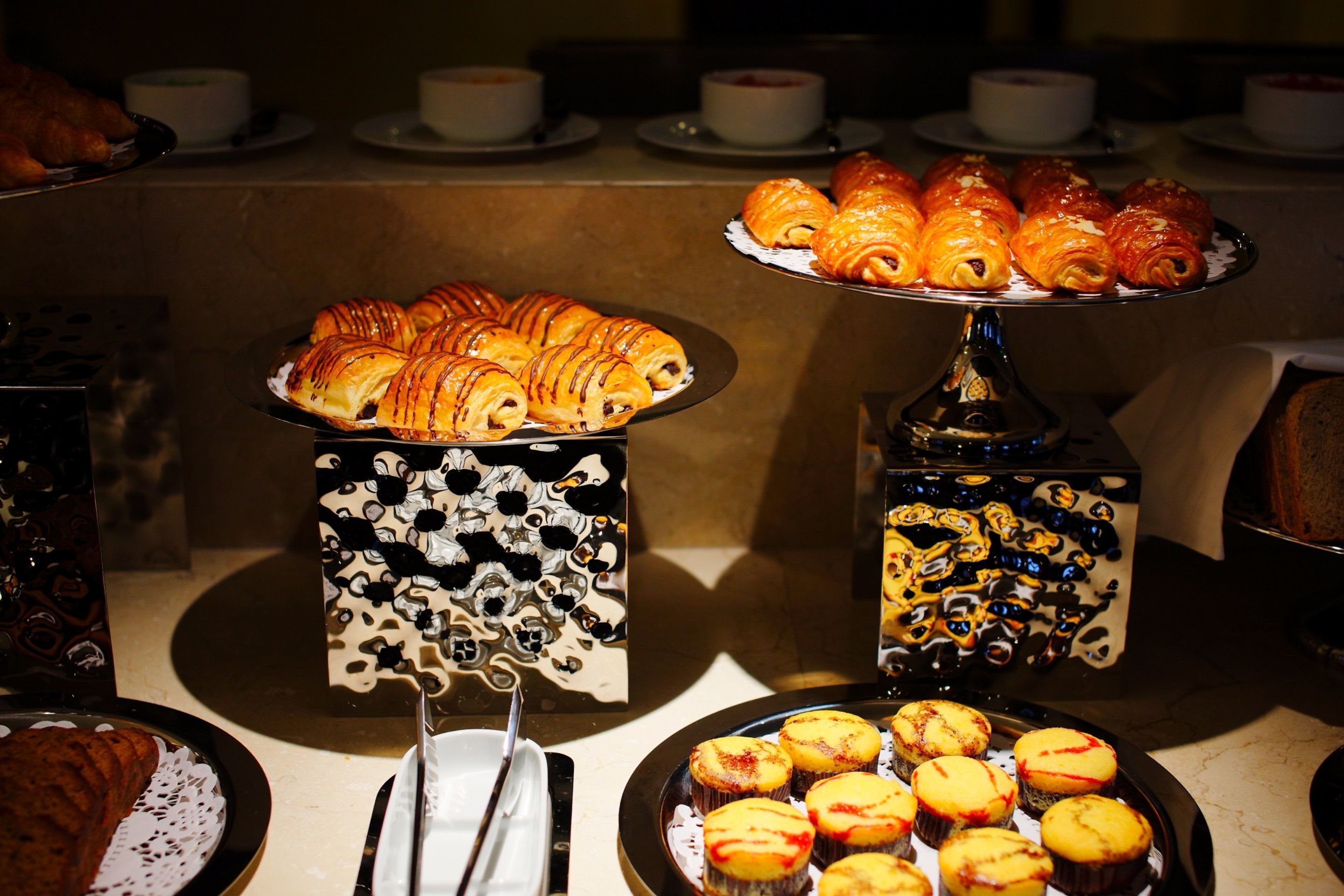 Un bufé con pastelería y productos de panadería, incluyendo croissants y muffins, dispuestos en elegantes soportes.
