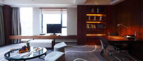 A modern hotel room with a flat-screen TV, a desk with a lamp, a bookshelf, and a dining area with a glass table and chairs.