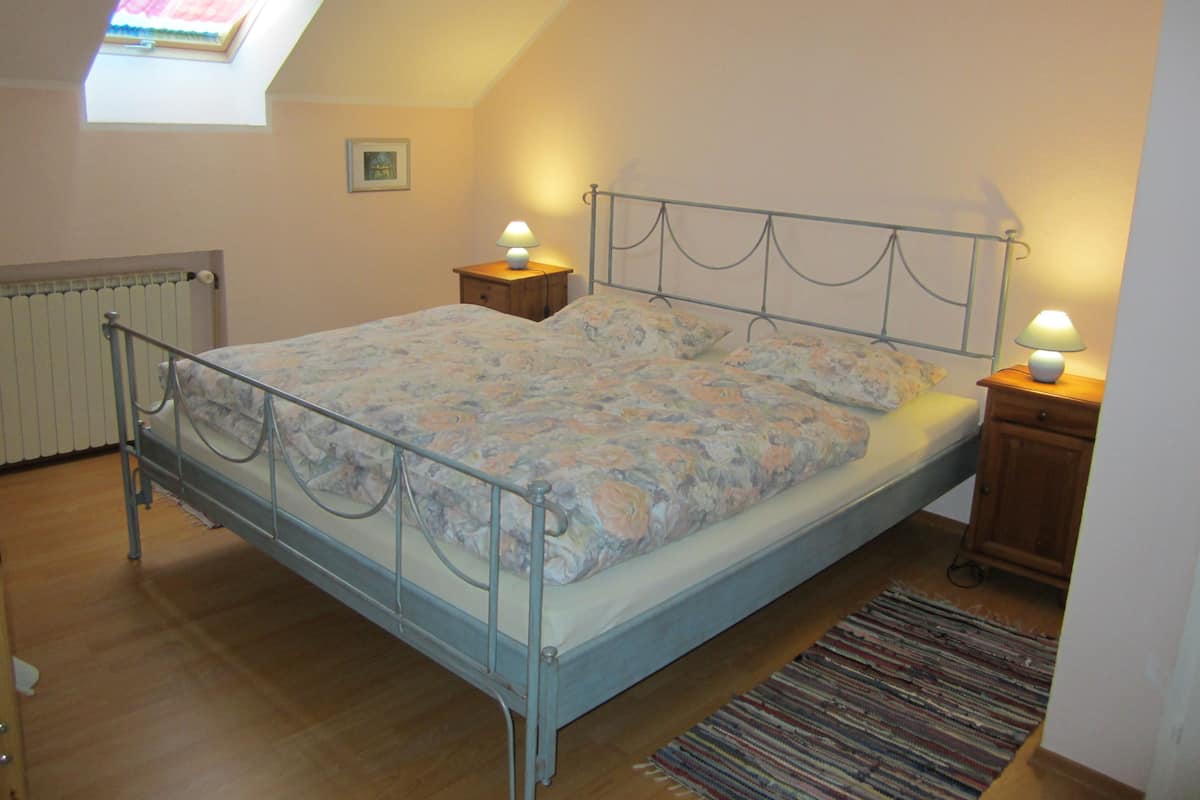 Een slaapkamer met een metalen bed, bloemenbedrukking, twee bedlampen, een radiator, een klein houten nachtkastje, een ingelijst schilderij aan de muur en een dakraam.