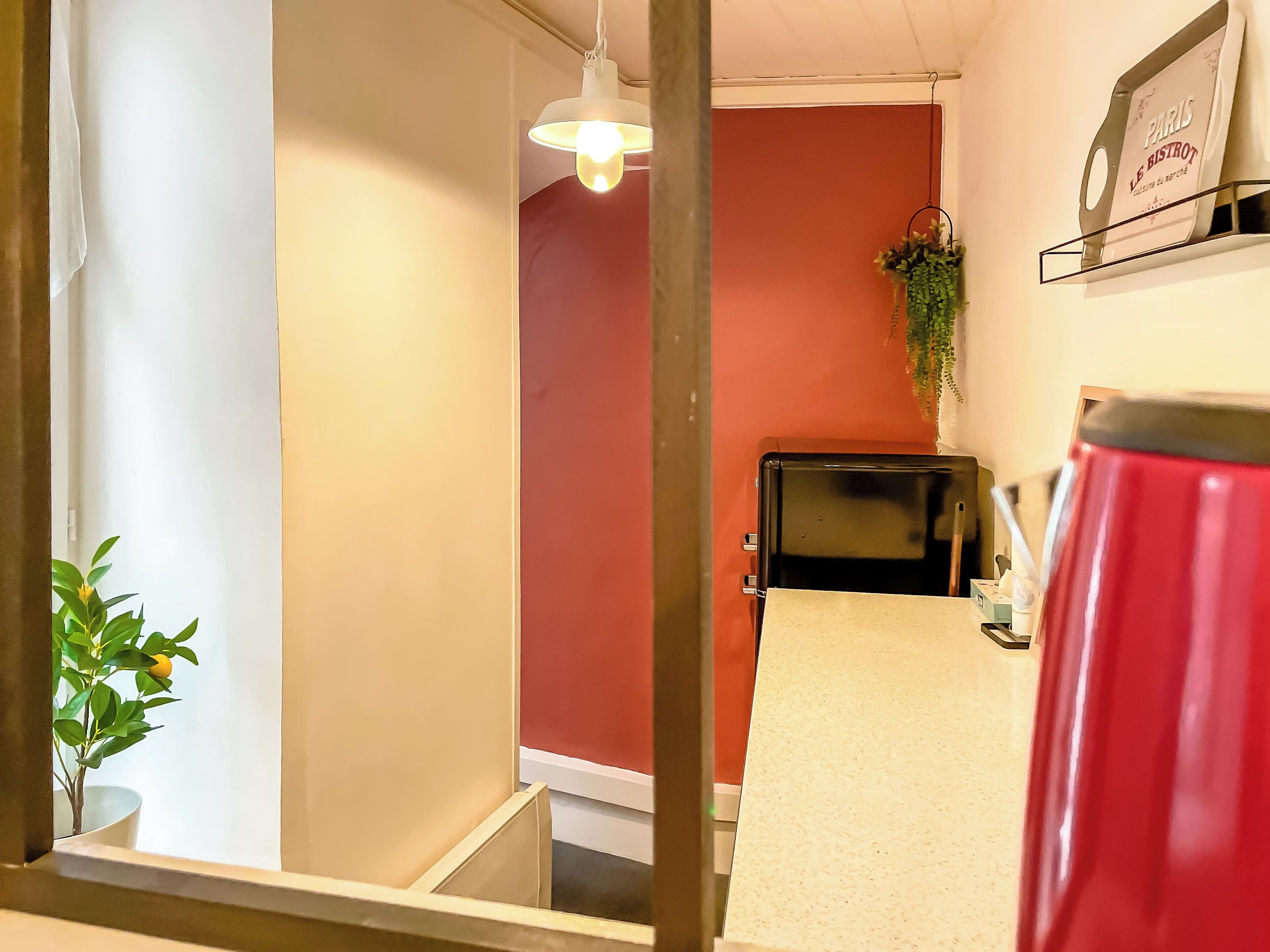 A hotel room with a red accent wall, a black desk, a white chair, a hanging plant, and a sign on the wall reading "Paris Le Bistro".