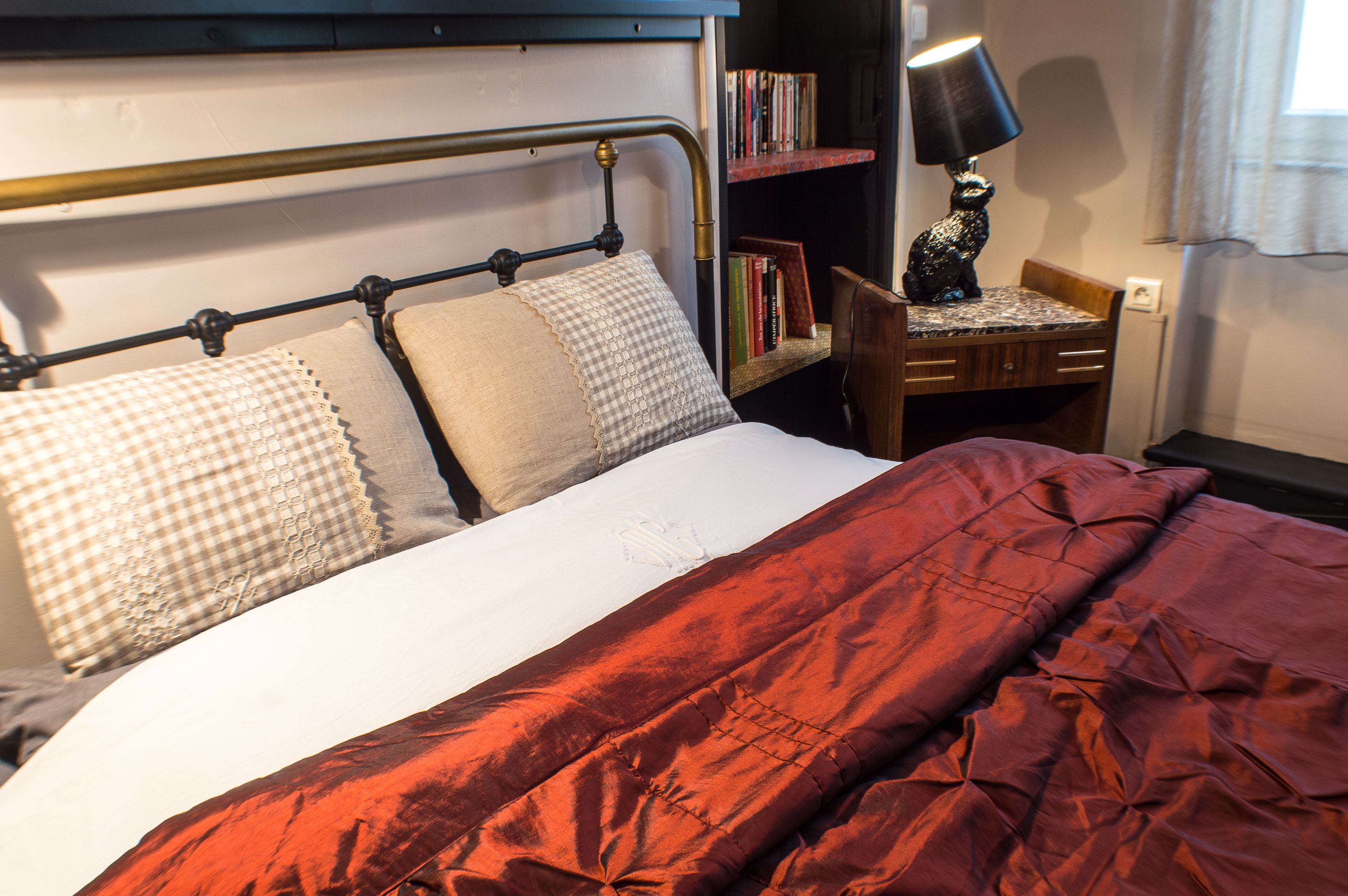 A neatly made bed with a red blanket, a checkered pillow, and a white pillow. There is a bookshelf with books and a lamp to the right.