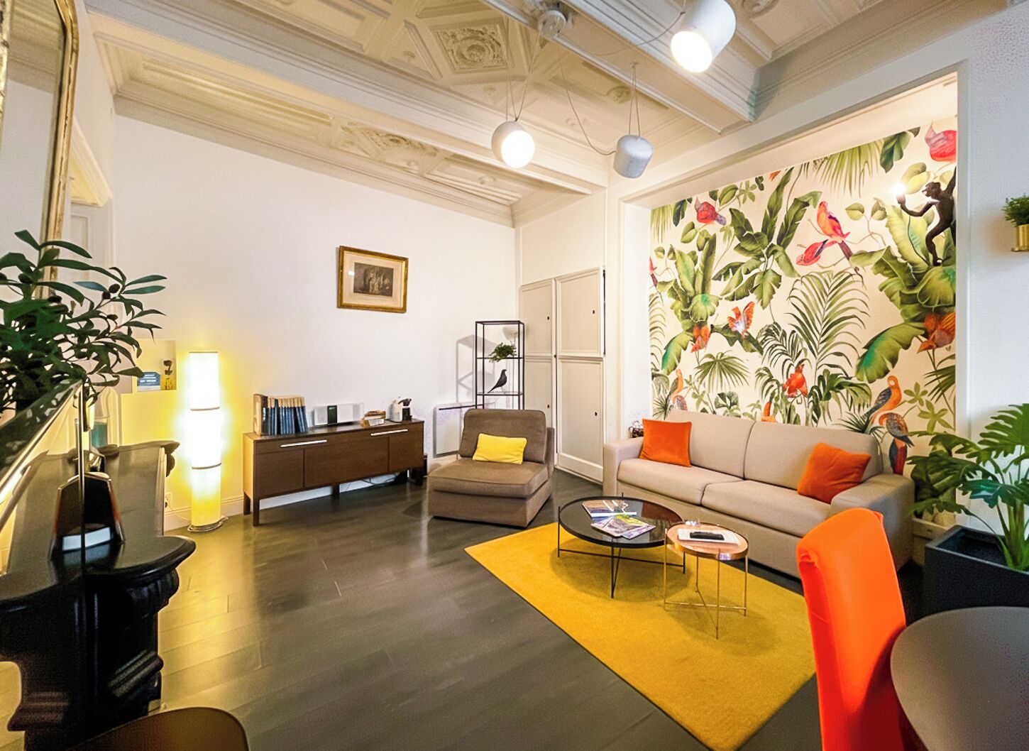 Een woonkamer met een bank, fauteuils, een salontafel, een boekenkast en een groot raam met tropische behang.