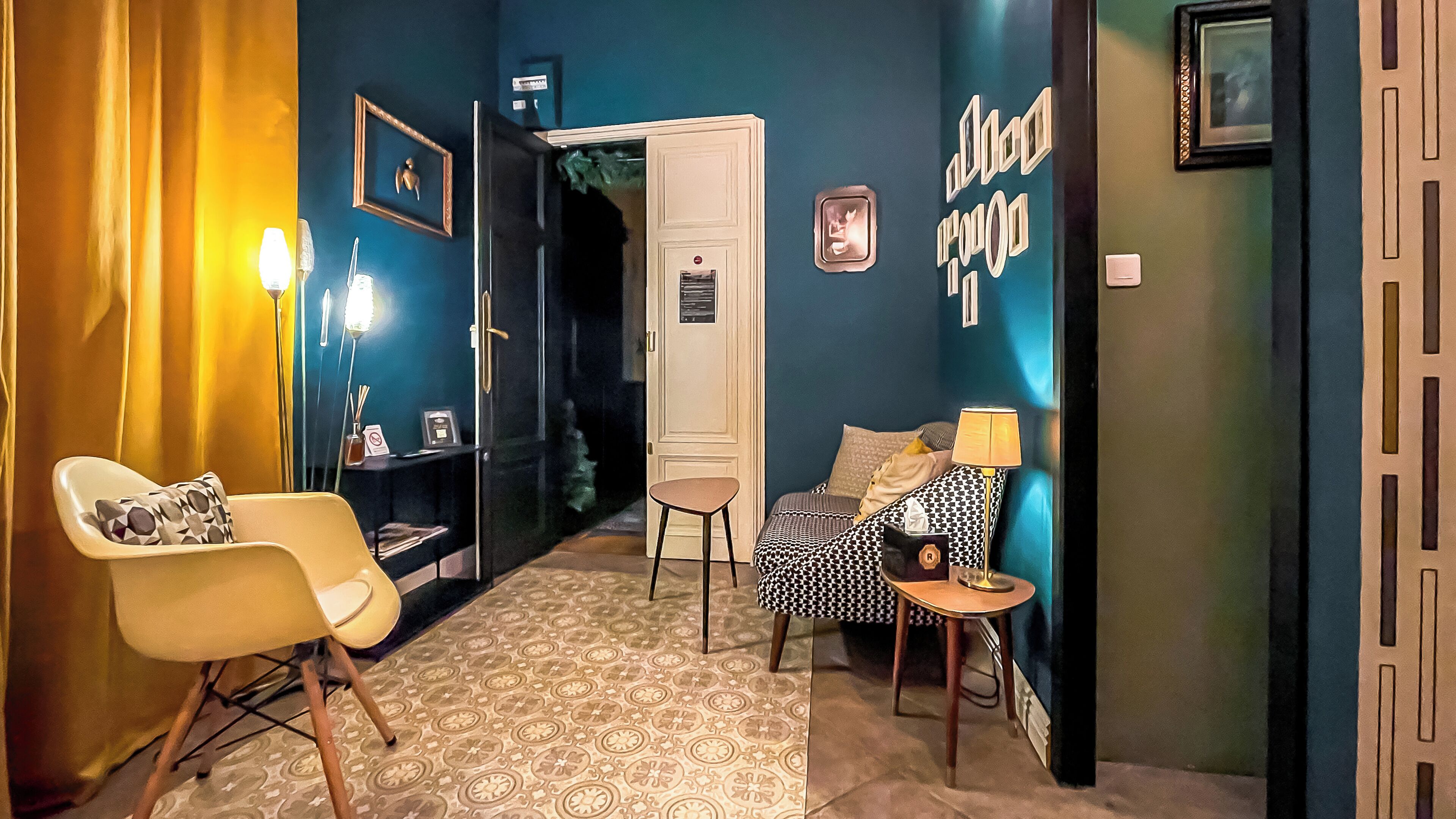 Una habitación con suelo estampado, un sillón blanco con un cojín de diseño geométrico, un sofá con estampado y una pequeña mesa con una lámpara.
