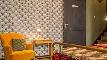 Una habitación con papel tapiz estampado, un sillón amarillo, una mesita de noche con una lámpara, una cama con una manta de piel y una puerta con la etiqueta 'Chambre'.