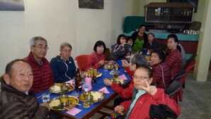 A group of people seated around a table with various dishes and drinks, in a room with a shelf and a television in the background. - Mystique Highland Resort (Pokhara)