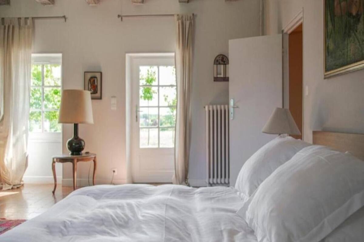 Une chambre avec un grand lit, deux fenêtres avec des rideaux, un radiateur et un tableau accroché au mur.
