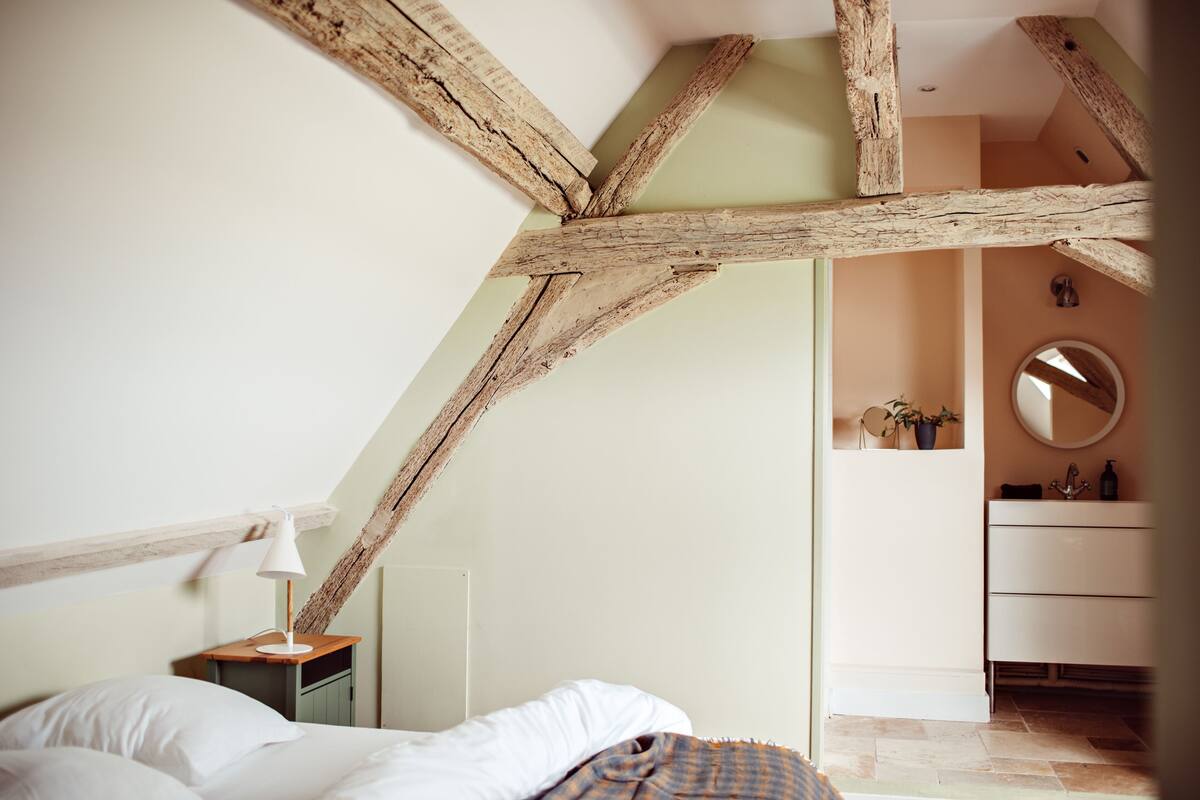 Une chambre avec des poutres apparentes, un lit recouvert de draps blancs, une table de chevet avec une lampe, et une salle de bain avec un miroir et un lavabo.
