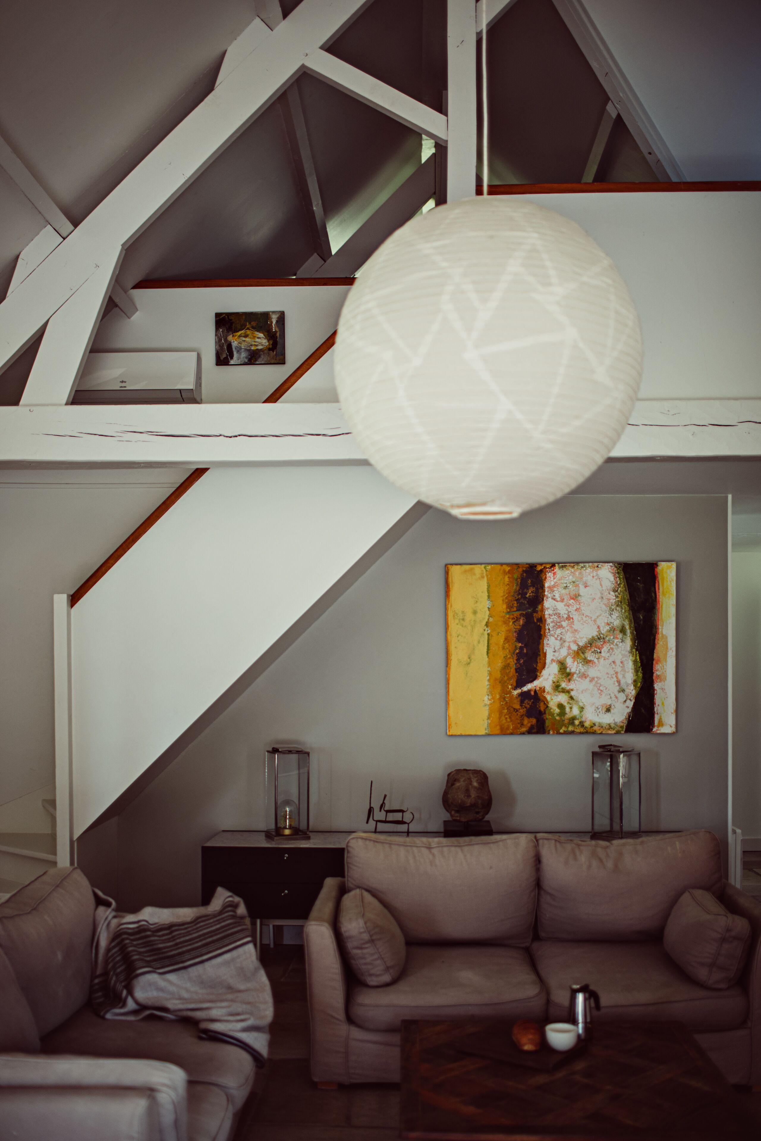 A loft-style living room with a high ceiling, exposed beams, a large abstract painting, a sofa, a coffee table, and a hanging light fixture.