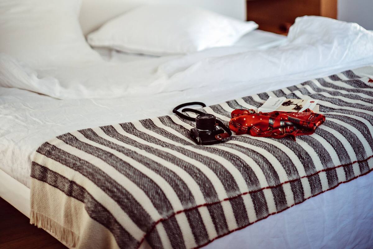 Un lit bien fait, recouvert d’une couverture à rayures et sur lequel est posé un sac rouge. Un poster illustré d’arbres est accroché au mur, au-dessus du lit.
