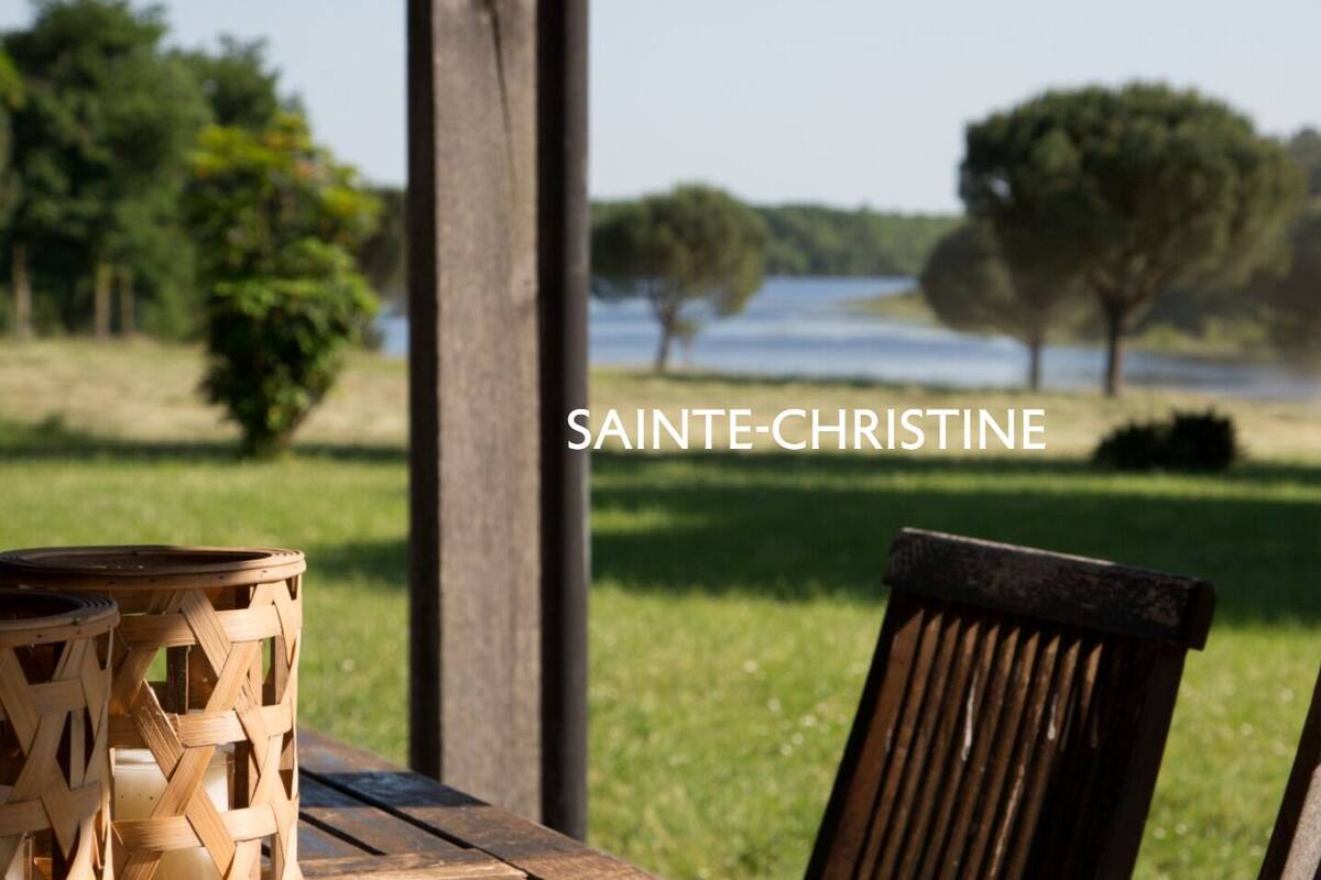 Une table et des chaises en bois, accompagnées d’un panier en osier, sont disposées de manière à offrir une vue sur une zone herbeuse, des arbres et une étendue d’eau.