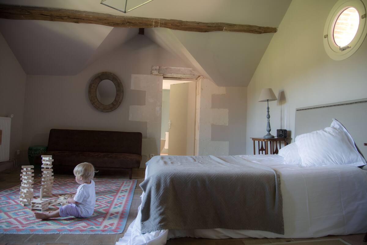 Un enfant joue avec des blocs de bois sur un tapis à motifs dans une pièce où se trouvent un lit, une lampe et une fenêtre ronde.